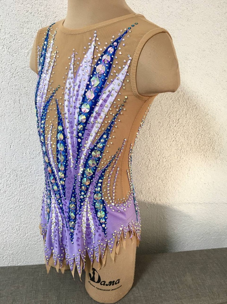 May include: A purple and blue leotard with a geometric design and rhinestones. The leotard has a scoop neck and sleeveless design. The leotard is on a dress form.