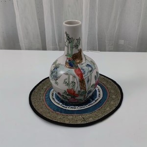 Puede incluir: Un jarrón de cerámica blanco con cuerpo redondo y cuello largo, decorado con pájaros coloridos, bambú y flores. El jarrón se encuentra sobre una alfombra redonda bordada con centro turquesa y ribete dorado.