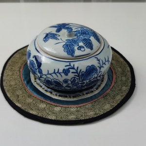 Puede incluir: Un tarro de cerámica blanco y azul con tapa, con diseños florales. El tarro se encuentra sobre una estera redonda decorativa con un patrón dorado y negro, y un borde turquesa y rosa. Estilo asiático tradicional.