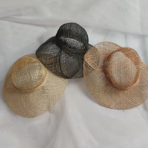 May include: Three straw hats in neutral tones. One is black, one is tan, and the other is a light beige. The hats have a woven texture and a rounded crown with a wide brim. The hats are arranged on a white surface.