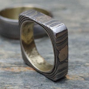May include: A square-shaped ring with a patterned, layered design in shades of grey and silver. The interior of the ring has a gold tone. Another ring is slightly out of focus in the background.