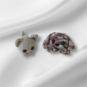 May include: Two small, crocheted animal figures. One is light gray with black eyes and pink accents, resembling a bunny. The other is brown and tan with black eyes, possibly a dog or bear. Both are handmade and have a soft, textured appearance.
