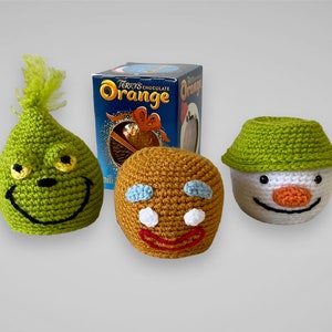 May include: Three crocheted stuffed toys: a green Grinch, a brown gingerbread man, and a white snowman with a green hat. The snowman is wearing a green hat and has a carrot nose. The gingerbread man has blue eyes and a red smile. The Grinch has yellow eyes and a green hat. The toys are sitting on a white background.