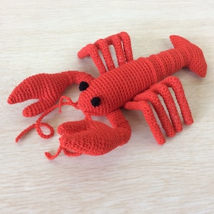 May include: A red crocheted lobster toy with black eyes. The lobster is lying on its back with its claws outstretched.