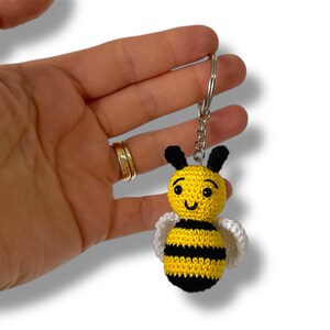 May include: A crocheted bee keyring. The bee is yellow with black stripes, black antennae, and white wings. The keyring is attached to a silver metal ring.