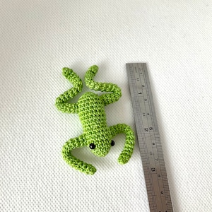 May include: A small green crocheted frog toy, approximately 2 inches in length, with black eyes. The frog is lying on a white surface with a ruler for scale.