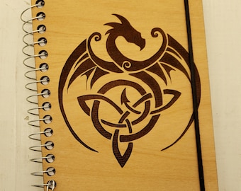 May include: A wooden spiral bound notebook with a black elastic closure. The cover features a laser-etched design of a dragon with Celtic knotwork.