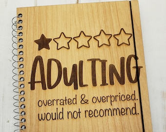 May include: A wooden spiral bound notebook with a black elastic closure. The cover is light brown wood with the text "ADULTING" in a large font. Below the text is a one-star rating with the text "overrated & overpriced. would not recommend."