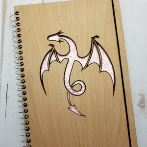 May include: A wooden spiral bound journal with a pink glitter dragon design cut out of the front cover.