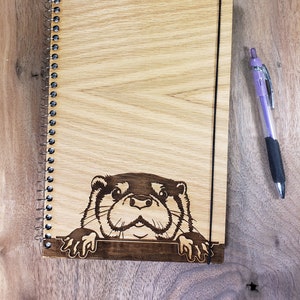 May include: A wood-grain spiral bound journal with a black elastic closure and a laser-etched image of an otter peeking over the top edge.
