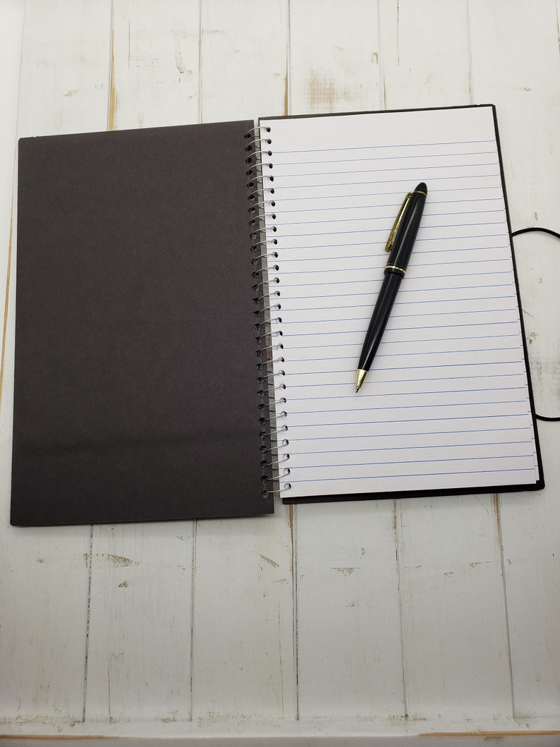 May include: A black and white spiral bound notebook with a black pen lying on the open page. The notebook is on a white wooden surface.