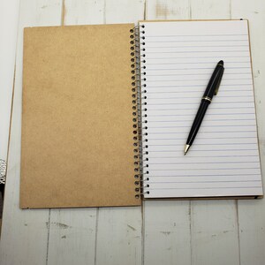 May include: A brown notebook with a spiral binding and lined pages. A black pen is lying on the open pages.