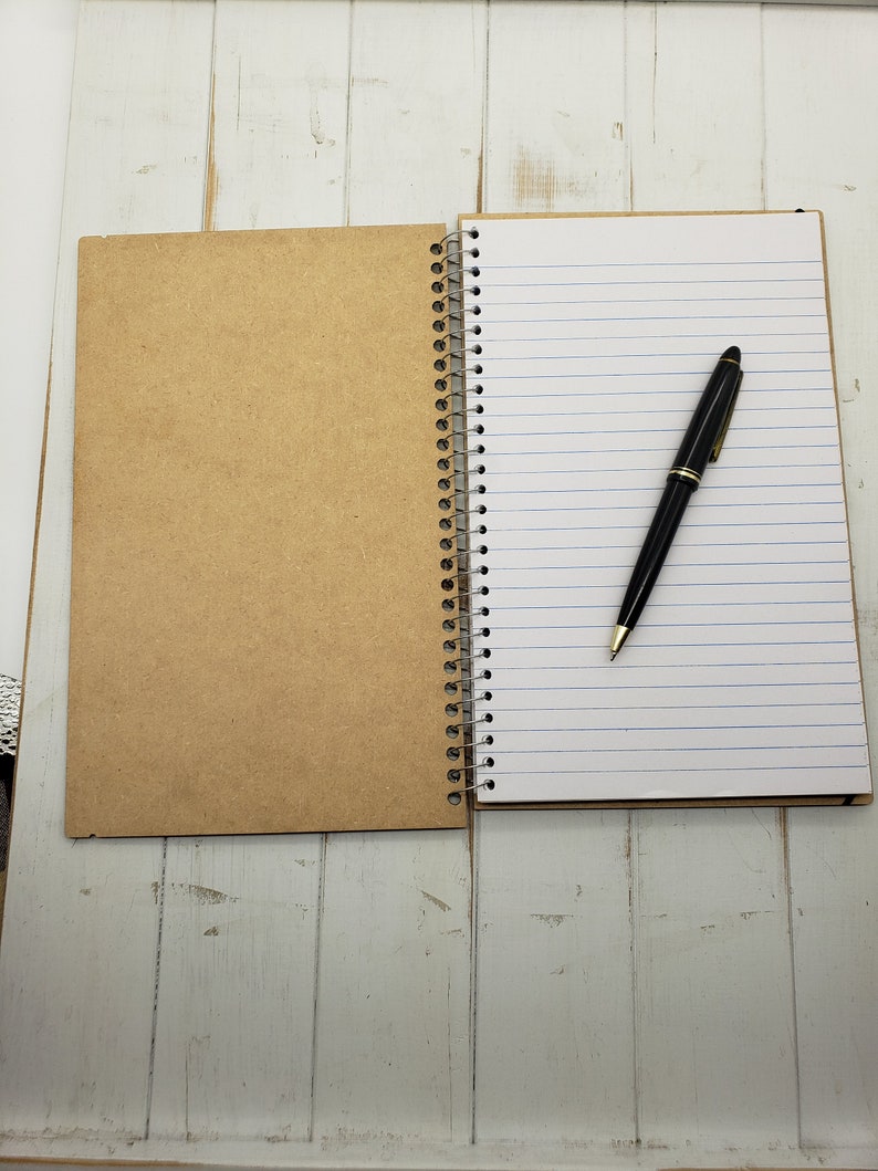 May include: A brown notebook with a spiral binding and lined pages. A black pen is lying on the open pages.