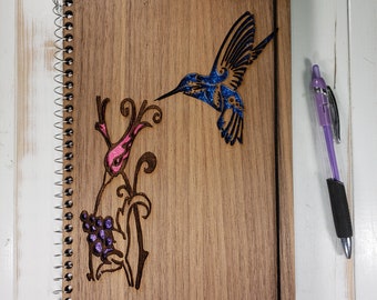 May include: A wooden journal with a spiral binding features a laser-cut design of a hummingbird and a flower with purple berries. The journal has a black ribbon bookmark.
