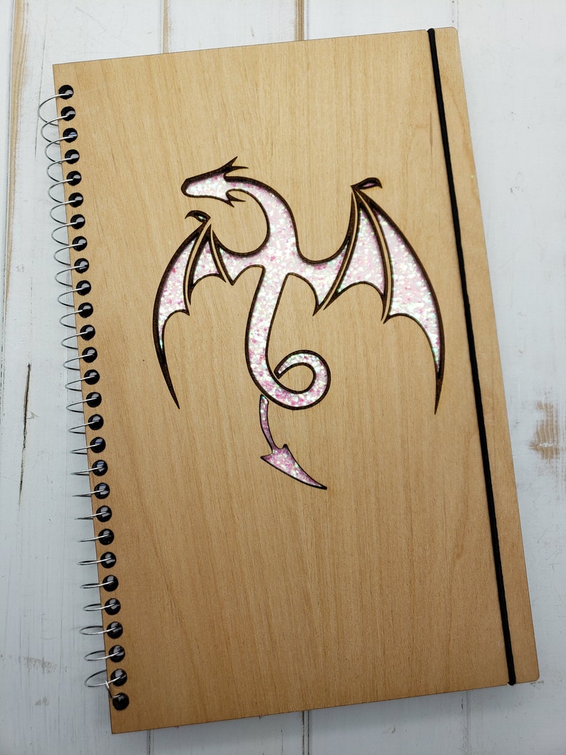 May include: A wooden spiral bound journal with a pink glitter dragon design cut out of the front cover.