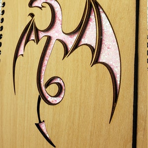 May include: A wooden journal with a pink glitter dragon design. The dragon is cut out of the wood and the glitter is in the cut-out area.