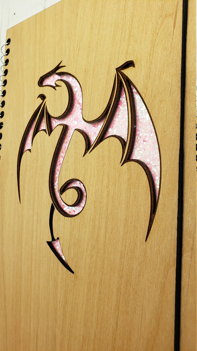 May include: A wooden journal with a pink glitter dragon design. The dragon is cut out of the wood and the glitter is in the cut-out area.