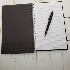 May include: A black and white spiral bound notebook with a black pen lying on the open page. The notebook is on a white wooden surface.
