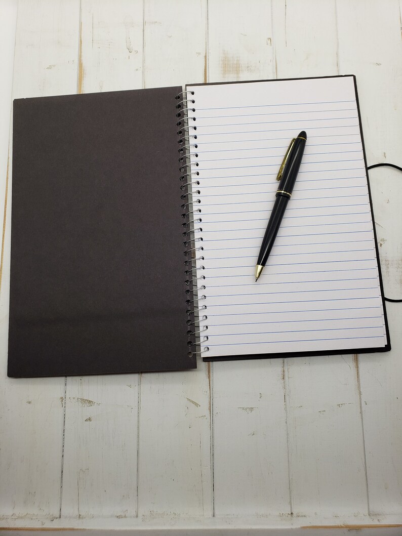 May include: A black and white spiral bound notebook with a black pen lying on the open page. The notebook is on a white wooden surface.