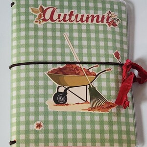 May include: A green and white checkered journal with a clear plastic cover. The front cover features a sticker of a wheelbarrow full of autumn leaves and a rake. The text "Autumn" is written in red on the cover.