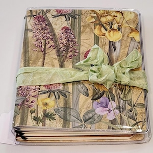 May include: A floral patterned journal with a green ribbon tied around it. The journal has a clear plastic cover and is closed.