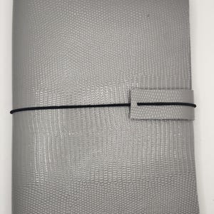 May include: A rectangular notebook cover in a light gray color with a textured, reptilian-like embossed pattern. The cover features a thin black elastic band that wraps around the exterior and secures through a matching gray tab on the front.