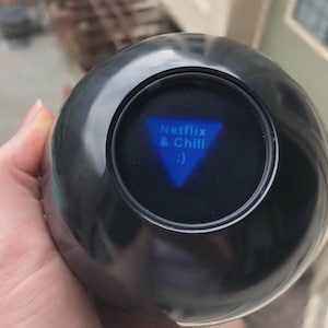 May include: A black fortune-telling ball with a blue triangular window displaying the text "Netflix & Chill :) ". The ball is held in a hand, with the background slightly blurred.