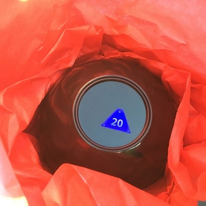 Pode incluir: Uma bola m&aacute;gica vermelha com um tri&acirc;ngulo azul e o n&uacute;mero 20 dentro. A bola est&aacute; embrulhada em papel de seda vermelho.