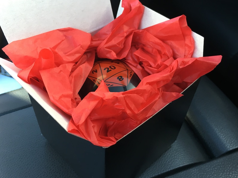 Pode incluir: Um dado preto e laranja de 20 faces em uma caixa preta com papel de seda vermelho.
