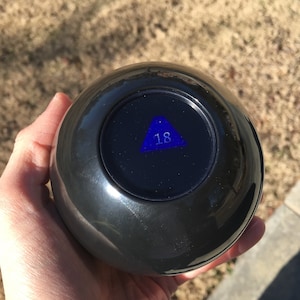 Pode incluir: Uma bola m&aacute;gica preta com um tri&acirc;ngulo azul e o n&uacute;mero 18 dentro.