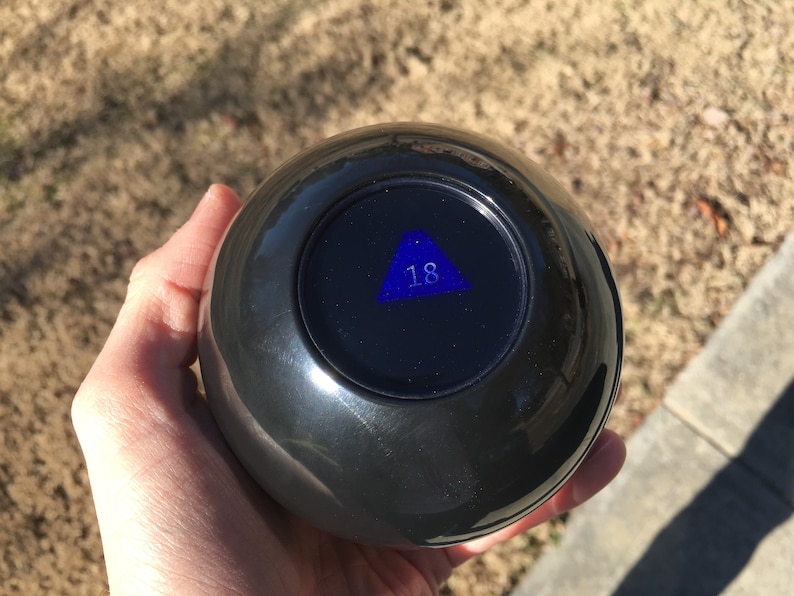Pode incluir: Uma bola m&aacute;gica preta com um tri&acirc;ngulo azul e o n&uacute;mero 18 dentro.