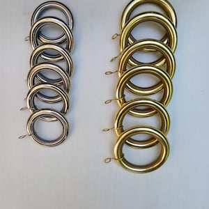 Puede incluir: Dos juegos de anillas para cortinas. Uno es plateado y el otro dorado. Cada anilla tiene un pequeño lazo metálico. Las anillas están dispuestas en dos pilas verticales sobre un fondo blanco.