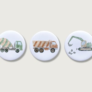 Könnte beinhalten: Drei weiße runde Buttons mit Aquarellillustrationen von Baumaschinen. Das Set enthält einen Betonmischer, einen Muldenkipper und einen Bagger, alle in Grün-, Braun- und Orangetönen.