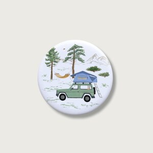 May include: A circular badge depicting a green SUV with a rooftop tent, set in a snowy scene with pine trees, a hammock, and mountains. The badge has a white background.