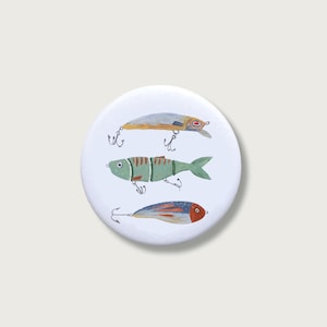 Könnte beinhalten: Ein runder weißer Button mit einer handgemalten Illustration von drei verschiedenen Angelködern. Der obere Köder ist silber und gold, der mittlere Köder ist ein segmentierter grüner Fisch und der untere Köder ist blau und rot gemustert.