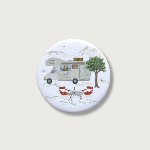 Könnte beinhalten: Ein weißer Button mit Camping-Motiv. Er zeigt ein graues Wohnmobil, das in der Nähe eines Baumes geparkt ist, mit roten Stühlen und einem Tisch zum Entspannen. Berge im Hintergrund erzeugen eine Szene voller Outdoor-Abenteuer und Reisen.