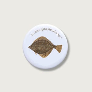 Könnte beinhalten: Ein runder, weißer Ansteckbutton mit einer detaillierten Illustration eines braun gesprenkelten Flundernfisches in der Mitte. Der Text "Du bist ganz flunderbar!" ist in einer dunkelgrauen serifenlosen Schrift bogenförmig über dem Fisch abgebildet.