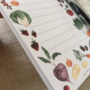 Puede incluir: Un bloc de notas blanco con una p&aacute;gina rayada y una variedad de ilustraciones de acuarela de verduras y frutas, incluyendo una remolacha roja, una cebolla amarilla, un tomate rojo, una col verde y fresas rojas. El bloc de notas tiene el texto "www.herzluft.de" impreso en la esquina inferior derecha.