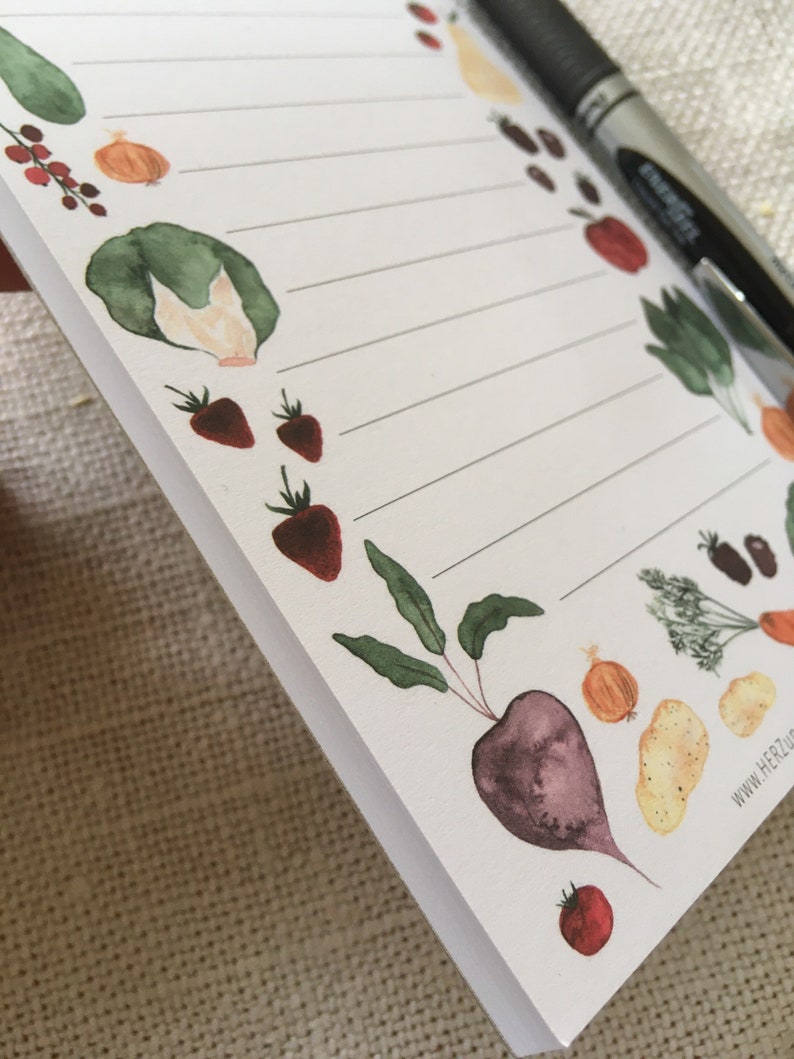 Puede incluir: Un bloc de notas blanco con una p&aacute;gina rayada y una variedad de ilustraciones de acuarela de verduras y frutas, incluyendo una remolacha roja, una cebolla amarilla, un tomate rojo, una col verde y fresas rojas. El bloc de notas tiene el texto "www.herzluft.de" impreso en la esquina inferior derecha.