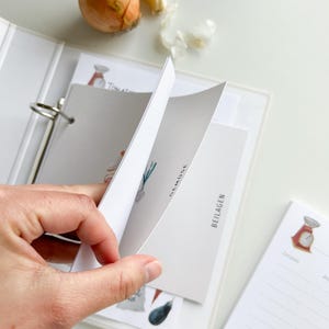 May include: A white binder with a clear plastic cover, holding recipe cards with illustrations of vegetables and the text "Gem&uuml;se" and "Beilagen". The binder has a ring mechanism and a white label on the spine. A notepad with a scale illustration and lined pages is visible on the right side of the image.
