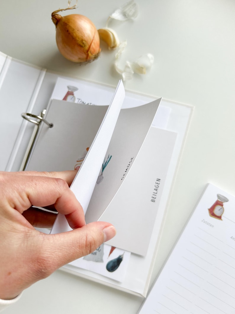 May include: A white binder with a clear plastic cover, holding recipe cards with illustrations of vegetables and the text "Gem&uuml;se" and "Beilagen". The binder has a ring mechanism and a white label on the spine. A notepad with a scale illustration and lined pages is visible on the right side of the image.