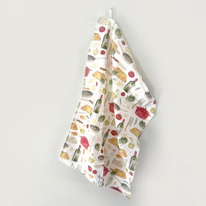 May include: White kitchen towel with a repeating pattern of cooking-related illustrations. The design includes red tomatoes, green wine bottles, pots, pans, and other kitchen utensils. The towel is hanging on a white wall.
