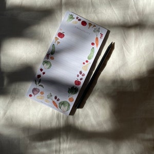 Puede incluir: Un bloc de notas blanco con un bol&iacute;grafo negro encima. El bloc de notas tiene un borde de ilustraciones coloridas de frutas y verduras. El texto "Einkaufsliste" est&aacute; impreso en el bloc de notas.