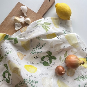 Op de afbeelding: Een witte linnen keukendoek met aquarel illustraties van kruiden, citroenen en knoflook. De doek heeft tekstlabels in verschillende talen. Een houten snijplank, een citroen en verse uien en knoflook zijn ook aanwezig.