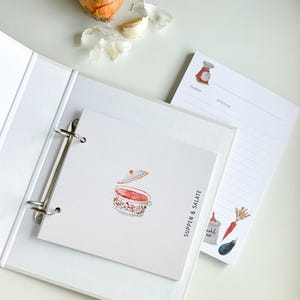 May include: A white binder with a ring mechanism and a white page with a watercolour illustration of a soup pot with a red lid and the text "Suppen & Salate" in black. The page is on top of a white page with a red scale illustration and the text "Zutaten" and "Anleitung" in black. There is a white page with a white and brown carrot, a brown aubergine, and a white bag with the text "mehl 405" in black.