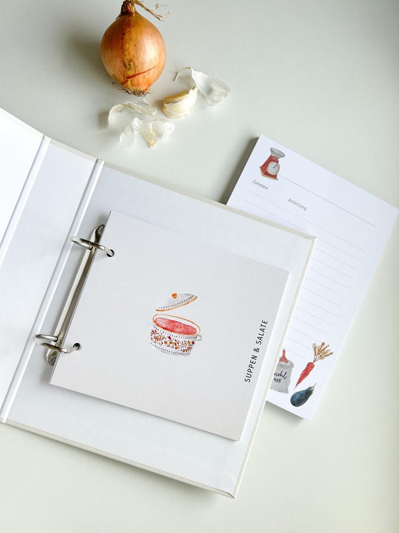 May include: A white binder with a ring mechanism and a white page with a watercolour illustration of a soup pot with a red lid and the text "Suppen & Salate" in black. The page is on top of a white page with a red scale illustration and the text "Zutaten" and "Anleitung" in black. There is a white page with a white and brown carrot, a brown aubergine, and a white bag with the text "mehl 405" in black.