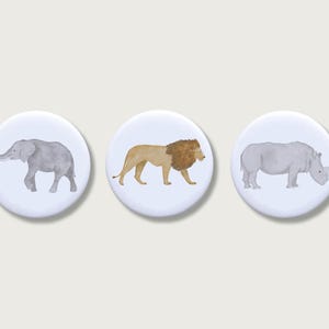 Könnte beinhalten: Drei runde, weiße Knöpfe mit Aquarellillustrationen afrikanischer Tiere. Die Knöpfe zeigen einen Elefanten, einen Löwen und ein Nashorn, jeweils in verschiedenen Grau- und Beigetönen. Die Knöpfe sind vor einem hellen Hintergrund platziert.