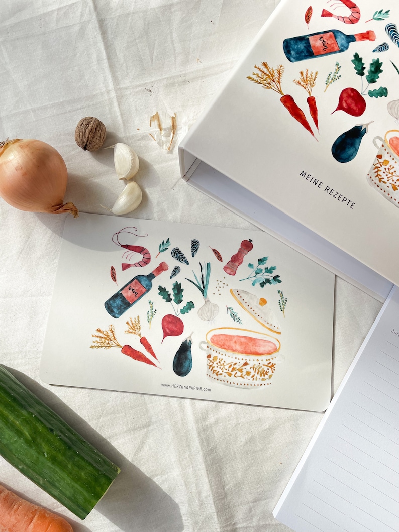 May include: A watercolour illustration of a pot of soup with various ingredients, including carrots, aubergine, garlic, wine, and herbs. The illustration is on a white card with the text "www.HERZundPAPIER.com" at the bottom. The card is on a white surface.