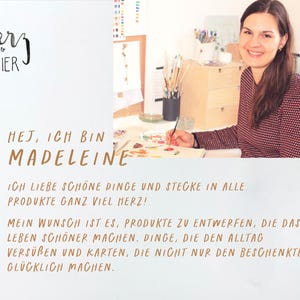 May include: A woman sits at a table with art supplies and paints. The text "HEJ, ICH BIN MADELEINE" is written in brown lettering. The text "ICH LIEBE SCH&Ouml;NE DINGE UND STECKE IN ALLE PRODUKTE GANZ VIEL HERZ!" is written in brown lettering. The text "MEIN WUNSCH IST ES, PRODUKTE ZU ENTWERFEN, DIE DAS LEBEN SCH&Ouml;NER MACHEN. DINGE, DIE DEN ALLTAG VERS&Uuml;BEN UND KARTEN, DIE NICHT NUR DEN BESCHENKTEN GL&Uuml;CKLICH MACHEN." is written in brown lettering.
