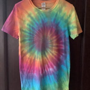 May include: A short-sleeved t-shirt with a vibrant tie-dye pattern. The shirt features a spiral design with a blend of colours including pink, orange, yellow, green, blue, and purple. The shirt is hanging on a wooden hanger.